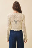 A woman viewed from the back wearing a cream-colored, long-sleeve crochet knit cropped top with a scalloped hem and dark blue jeans, set against a solid beige background.
