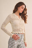 A woman wearing a cream-colored, long-sleeve crochet cardigan with a scalloped hem and button details, paired with light floral pants, set against a plain beige background.