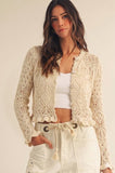 A woman wearing a cream-colored, long-sleeve crochet knit cardigan over a white crop top and white drawstring shorts, standing against a plain light beige background.