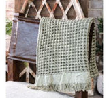 A sage green waffle fringe throw draped over the back of a chair, with a wooden lattice fence in the background.