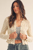A woman wearing a cream-colored, long-sleeve crochet cardigan over a gray tank top and blue jeans, posing against a plain beige background.
